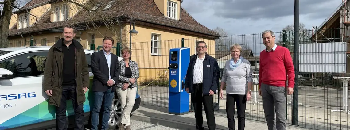 Mehrere lächelnde Menschen haben sich um eine EMB Ladestation für E-Mobilität versammelt. Foto: GASAG Solution Plus