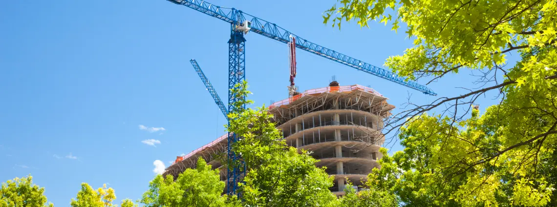 Ein Neubau mit Kränen, im Vordergrund grüne Bäume, im Hintergrund blauer Himmel